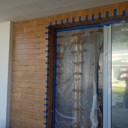 Angelika Krause - Montaż płytek imitujących drewno na ścianie przy drzwiach, zabezpieczonych folią. Widoczne niebieskie taśmy i krzyżyki dystansowe.