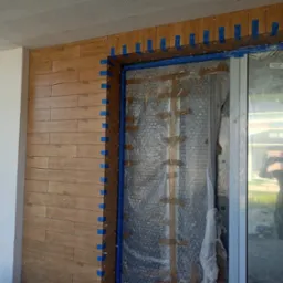 Montaż płytek imitujących drewno na ścianie przy drzwiach, zabezpieczonych folią. Widoczne niebieskie taśmy i krzyżyki dystansowe.