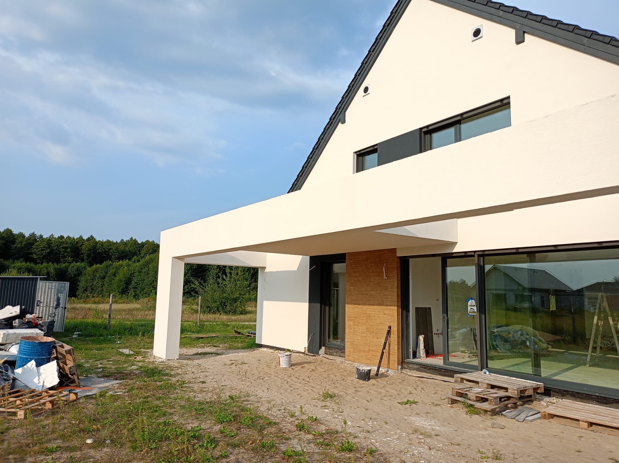 Nowoczesny dom jednorodzinny w trakcie wykończenia elewacji, z widocznymi materiałami budowlanymi i narzędziami na zewnątrz. Jasna fasada i duże przeszklenia.