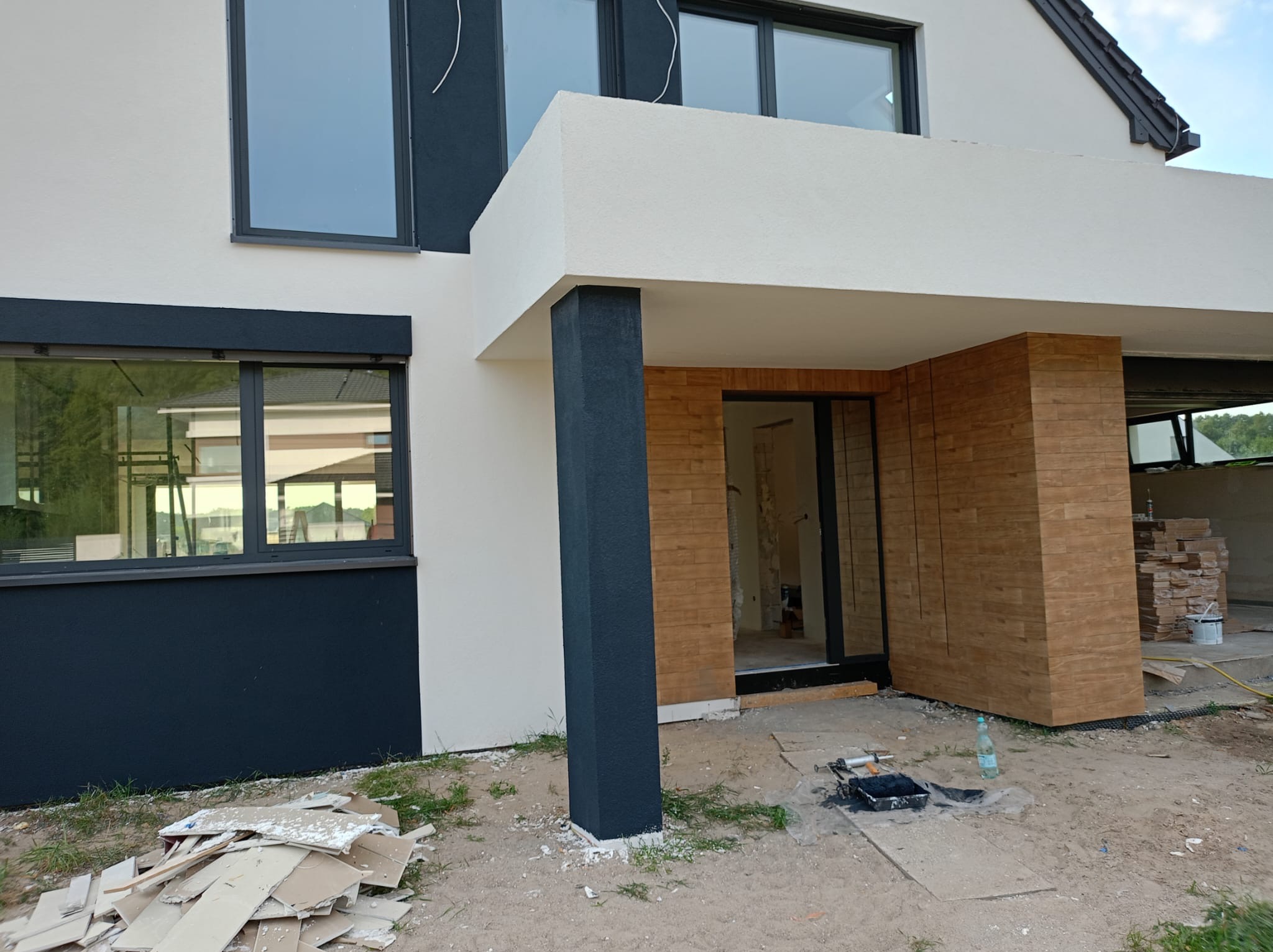 Elewacja domu jednorodzinnego w trakcie prac wykończeniowych. Widoczne pomalowane elementy, okładzina drewnopodobna i narzędzia malarskie.