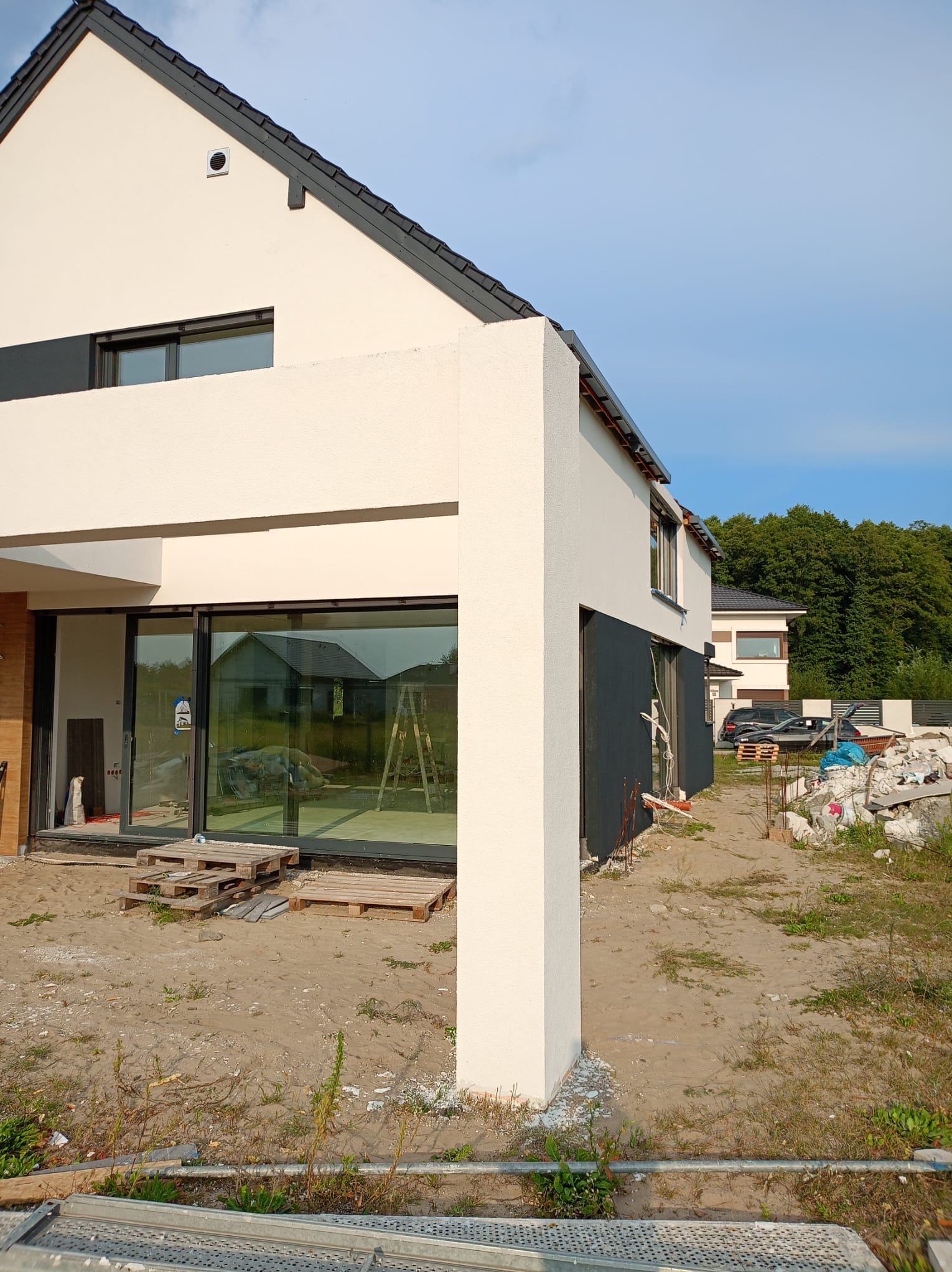 Elewacja domu jednorodzinnego w trakcie wykończenia, widoczne palety, rusztowania i gruz. Dom z nowoczesną architekturą, jasna elewacja z grafitowymi akcentami.