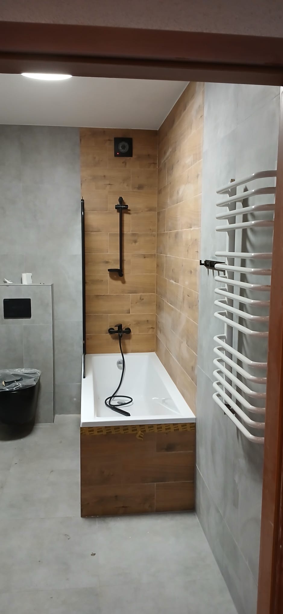 Wykończona łazienka z wanną obudowaną drewnopodobnymi płytkami, szara ściana, biały grzejnik drabinkowy i czarna armatura. Widok zza framugi drzwi.