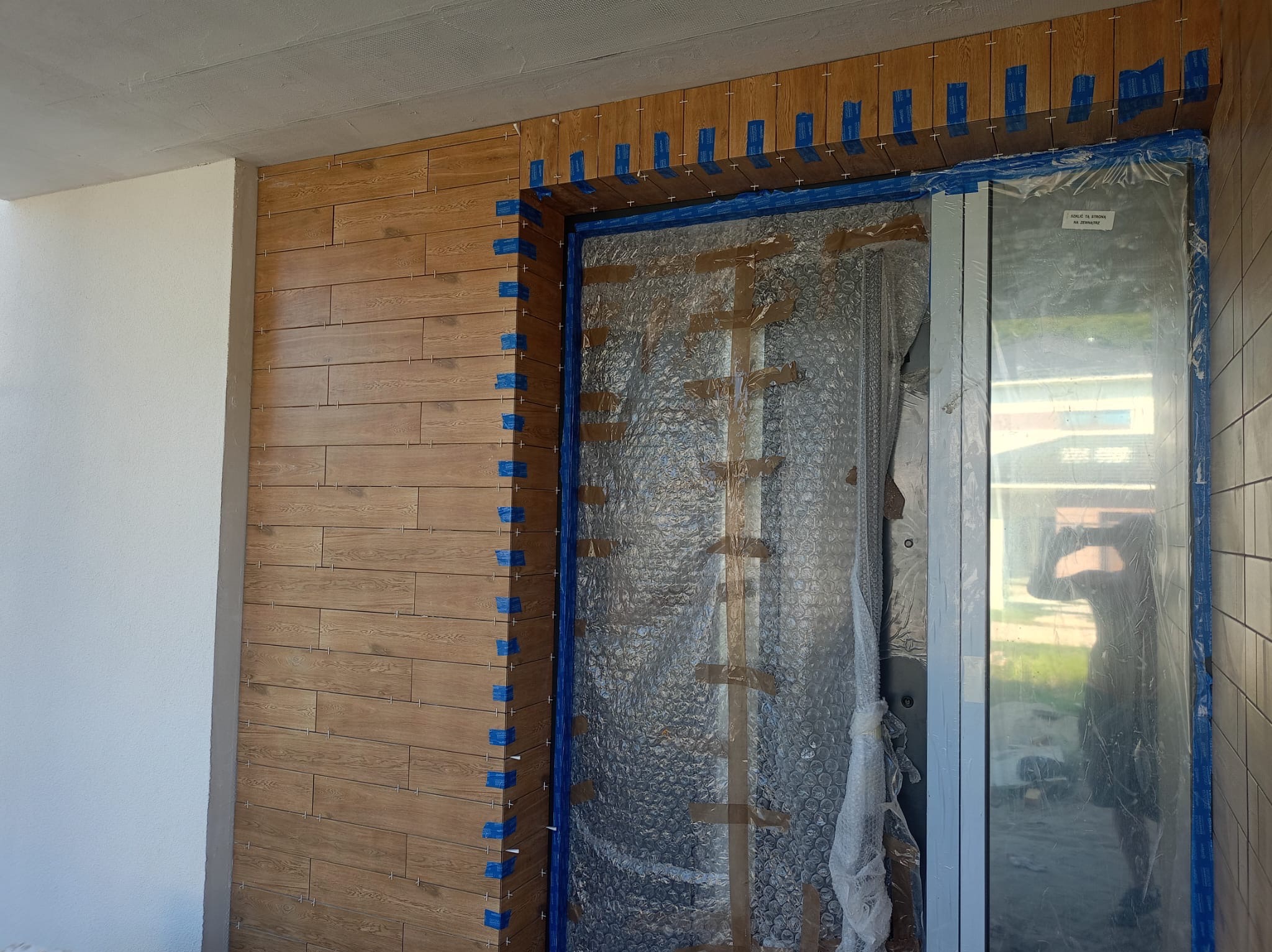 Montaż płytek imitujących drewno na ścianie przy drzwiach, zabezpieczonych folią. Widoczne niebieskie taśmy i krzyżyki dystansowe.