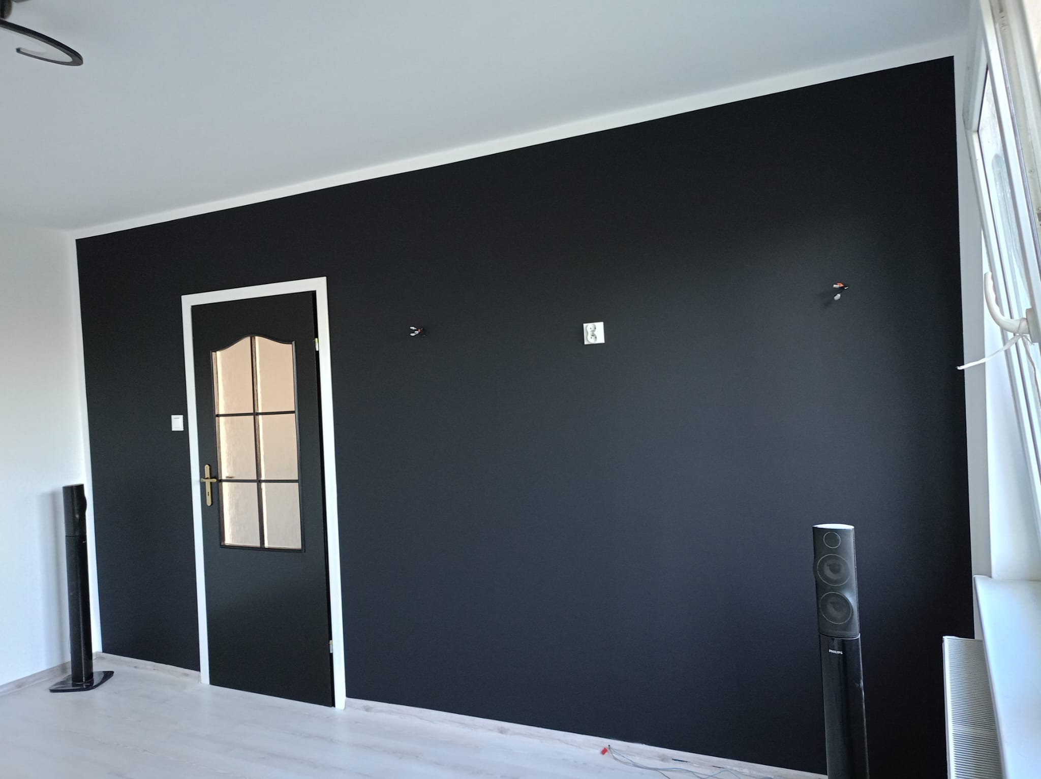Ściana pomalowana na czarno z białą stolarką drzwiową i przypodłogową. Widoczne głośniki pod ścianą. Minimalistyczny design wnętrza po malowaniu.