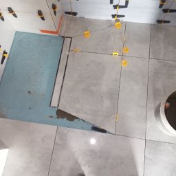 Ados.Usługi glazurnicze. - Układanie szarych płytek w łazience z odpływem liniowym. Widoczne klipsy poziomujące i buty robocze. Etap prac przed fugowaniem.