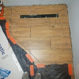 Ados.Usługi glazurnicze. - Ułożone drewnopodobne płytki w łazience z czarnym odpływem liniowym. Widoczne krzyżyki dystansowe i folia ochronna na podłodze. Buty osoby robiącej zdjęcie.