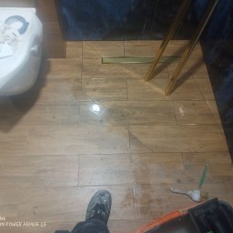 Ados.Usługi glazurnicze. - Ułożone drewnopodobne płytki w łazience z odpływem liniowym. Widoczne narzędzia glazurnicze i fragment toalety. Płytki ułożone w jodełkę przy odpływie.