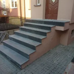 Zewnętrzne schody wejściowe wyłożone szarym kamieniem, prowadzące do brązowych drzwi. Elewacja budynku w odcieniach beżu. Widok na bruk i fragment ogrodu.