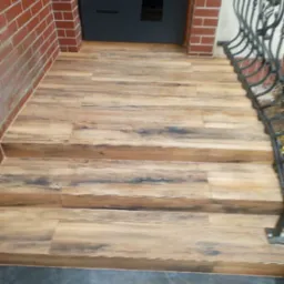 Schody wejściowe wyłożone panelami imitującymi drewno, z ciemnymi akcentami, prowadzące do czarnych drzwi. Po prawej stronie widoczna metalowa balustrada.