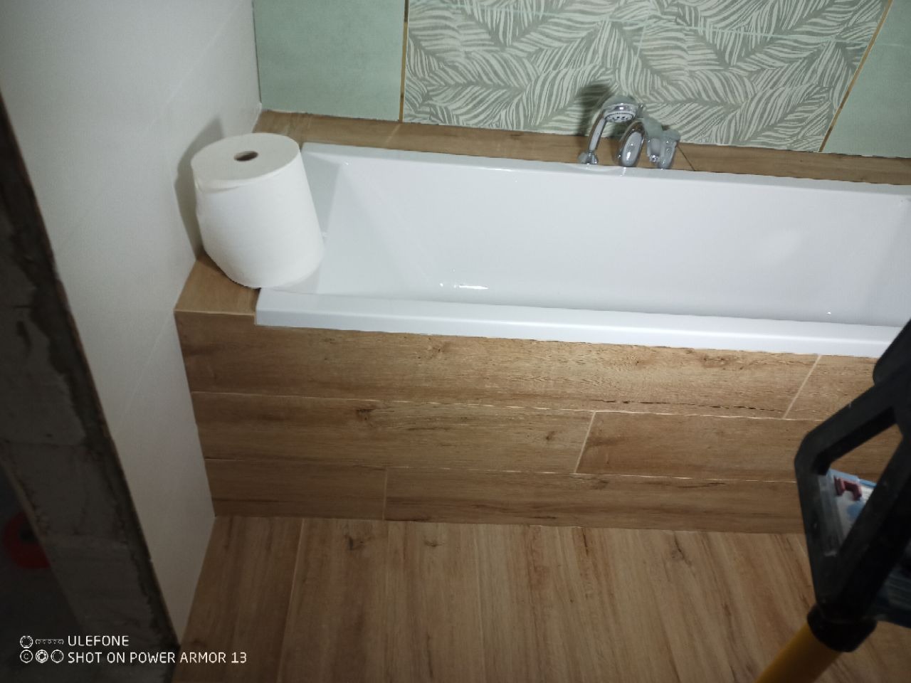 Wykończona łazienka z wanną obudowaną płytkami imitującymi drewno, z rolką papieru toaletowego i baterią. Na ścianie płytki w liście.