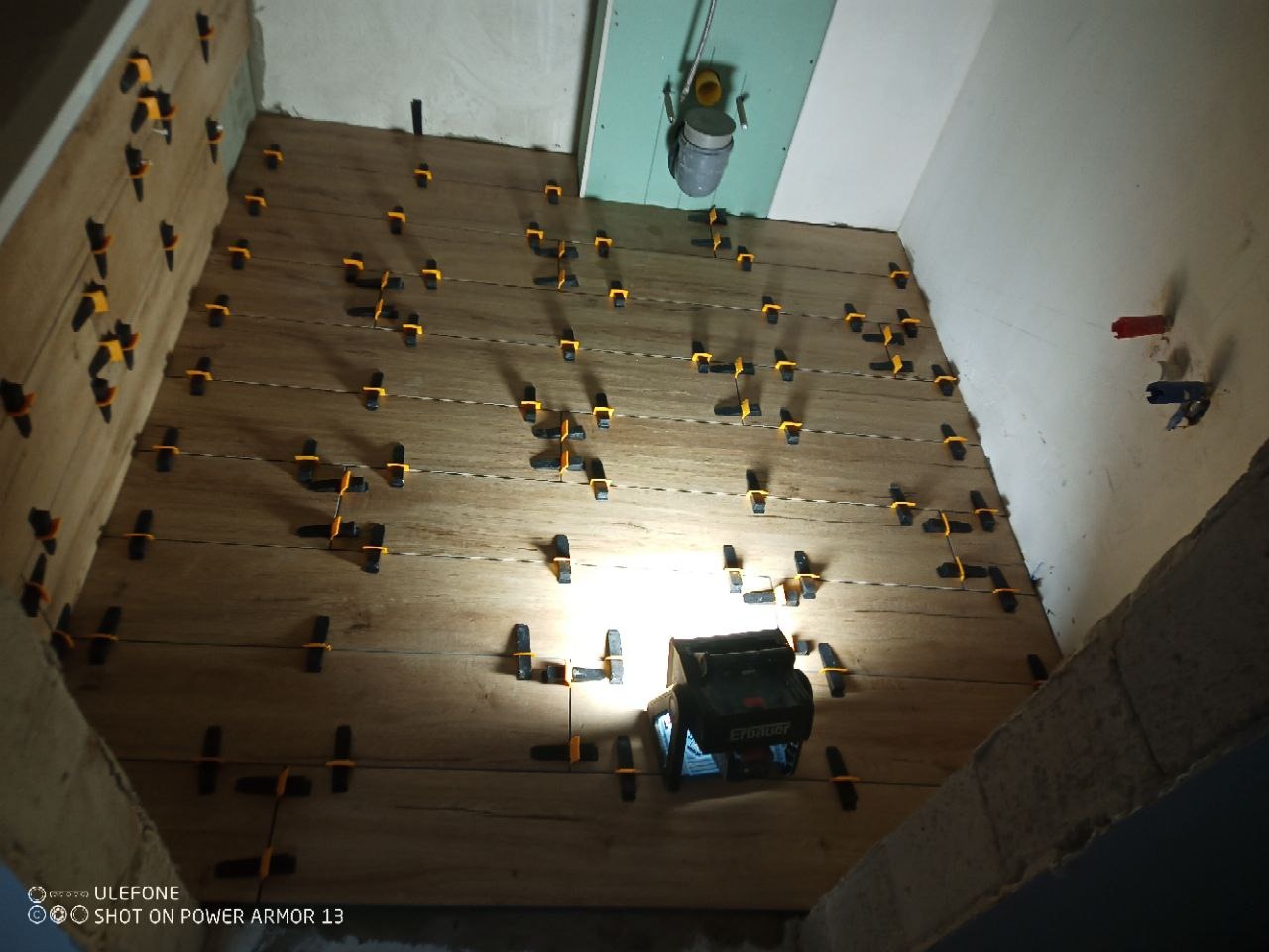 Układanie drewnopodobnych płytek z użyciem poziomicy laserowej i klipsów poziomujących w trakcie remontu. Widoczna płyta karton gips.