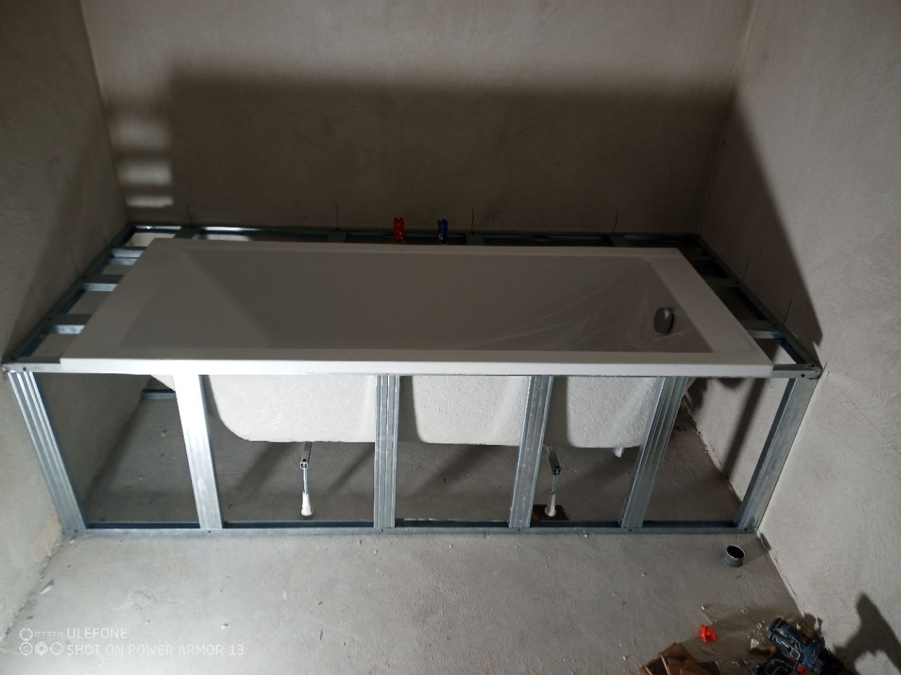 Biała wanna osadzona w metalowej konstrukcji w surowej łazience, widoczne rury i elementy instalacji, przygotowanie do obudowy wanny, szare ściany w tle.