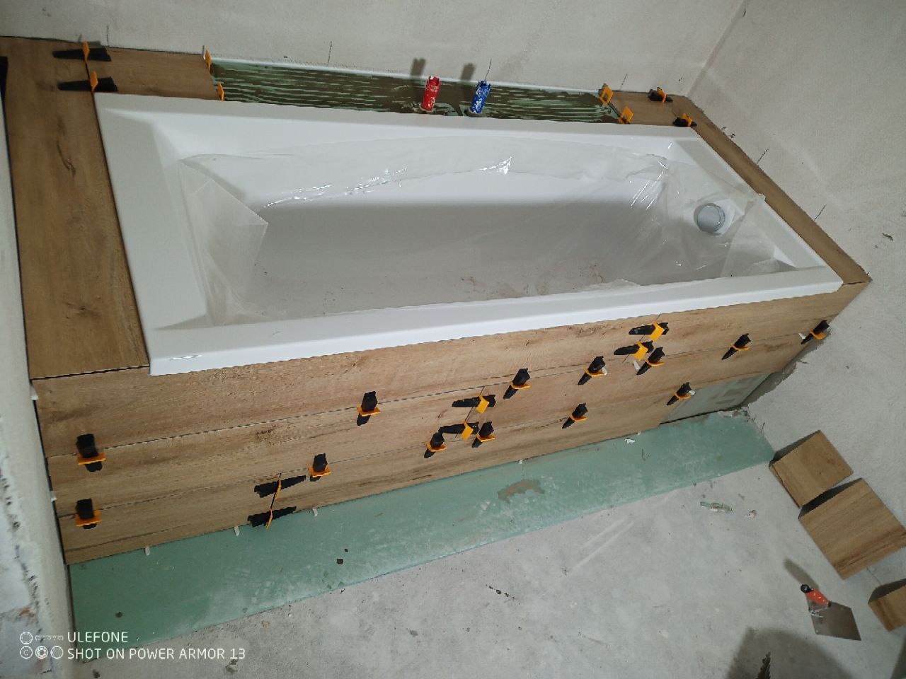 Obudowa wanny z płytek imitujących drewno, z użyciem klipsów poziomujących. Widoczna biała wanna zabezpieczona folią. Nierówne tło surowej ściany i podłogi.