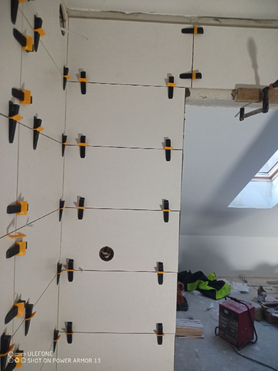 Białe płytki ścienne z czarno-żółtymi klipsami poziomującymi, czarne sznurki, widok na wnętrze remontowanego poddasza z narzędziami i oknem dachowym.