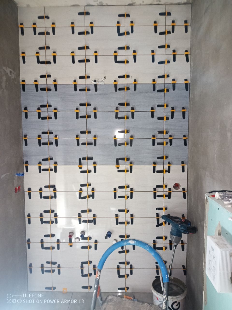 Ściana łazienki w trakcie układania płytek z wzorem geometrycznym, używane są klipsy poziomujące. Widoczna drabina i narzędzia glazurnicze.