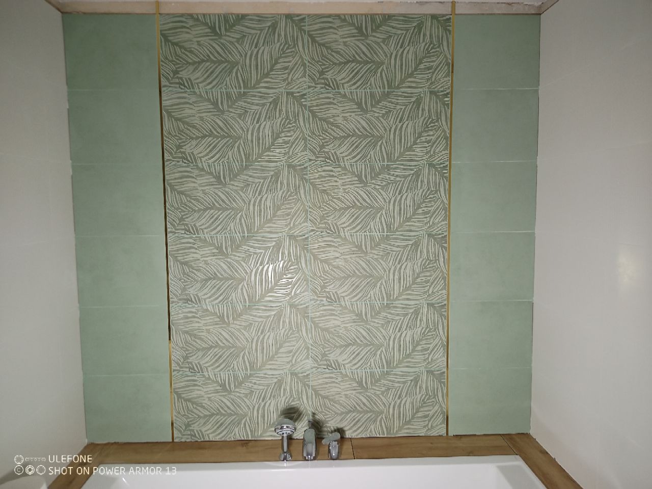 Wykończona ściana nad wanną z zielonymi płytkami. Centralna część zdobiona wzorem liści, obramowana listwami i gładkimi płytkami. Widoczne baterie umywalkowe.