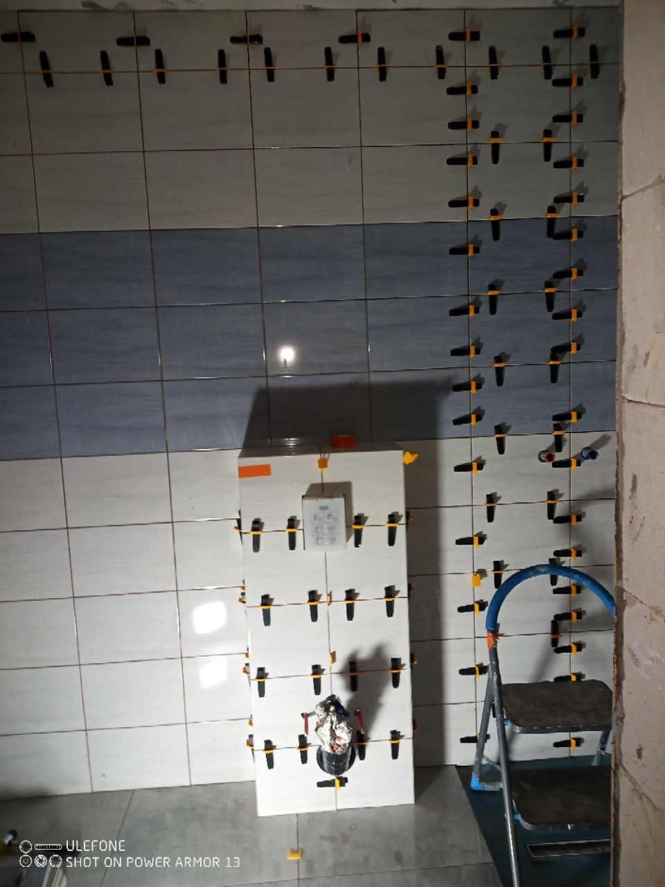 Ściana w trakcie układania płytek w łazience, z użyciem klipsów poziomujących. Widoczna drabina i instalacja pod umywalkę. Część płytek w kolorze niebieskim.