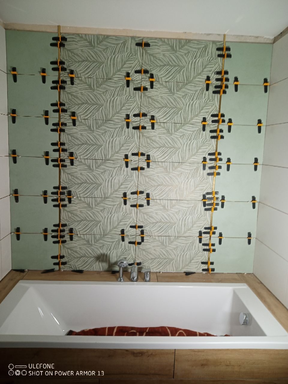 Układanie płytek z motywem liści w łazience w Słupsku. Widoczne systemy poziomowania glazury i fragment wanny. Etap prac wykończeniowych.