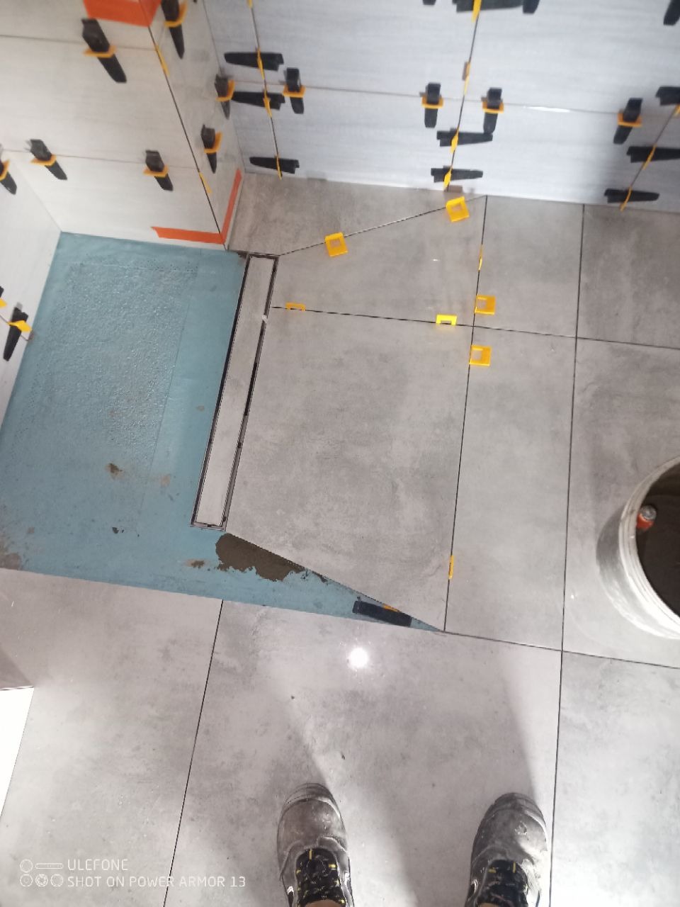 Układanie szarych płytek w łazience z odpływem liniowym. Widoczne klipsy poziomujące i buty robocze. Etap prac przed fugowaniem.