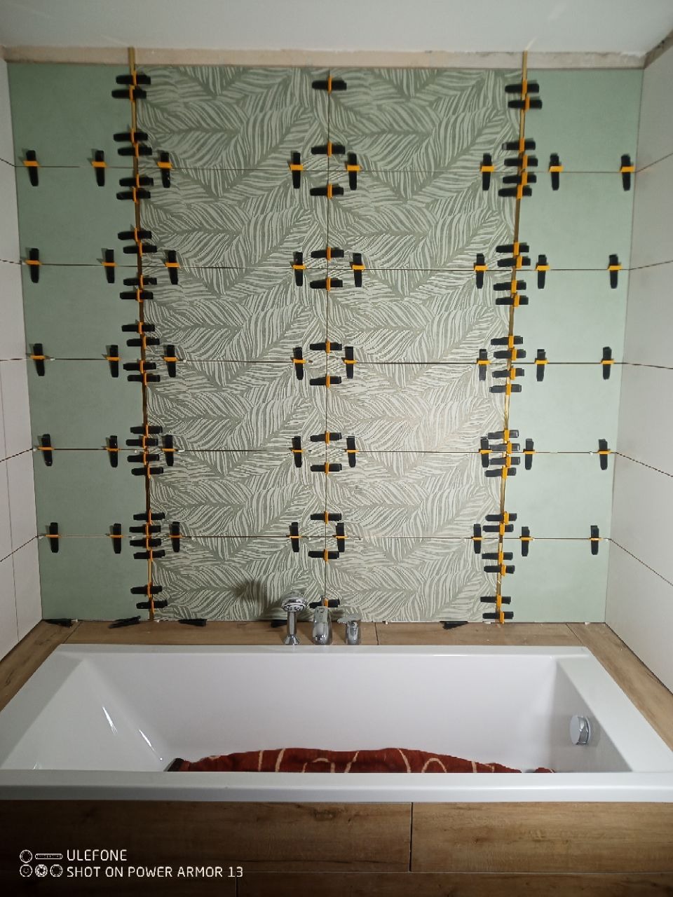 Łazienka w trakcie remontu: ułożone płytki z motywem liści, krzyżyki dystansowe, wanna. Widoczne etapy prac glazurniczych, dbałość o wzór i precyzję układania.