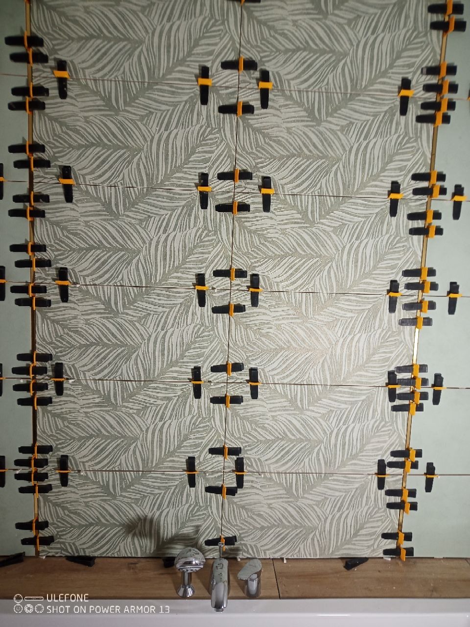 Układanie płytek z motywem liści na ścianie. Widoczne klipsy poziomujące. Ujęcie z bliska na fragment ściany w trakcie prac glazurniczych.