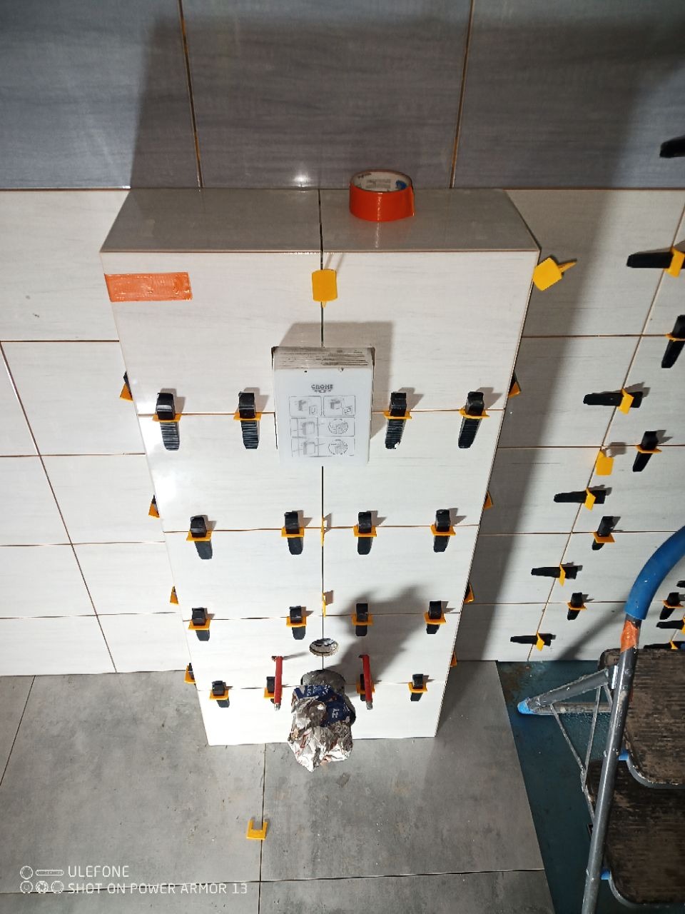 Białe płytki ścienne z czarnymi klipsami poziomującymi, pomarańczowa taśma, instrukcja Grohe i zabezpieczona folią instalacja wodna w trakcie prac glazurniczych.