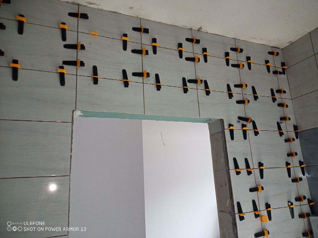 Świeżo ułożone, szare płytki ścienne w łazience, z użyciem czarnych klipsów i żółtych klinów poziomujących, widoczne krawędzie otworu drzwiowego.