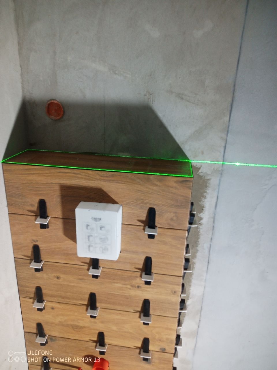 Układanie drewnopodobnych płytek z użyciem lasera i klipsów poziomujących. Widoczny panel sterowania Grohe. Surowy beton na ścianie.