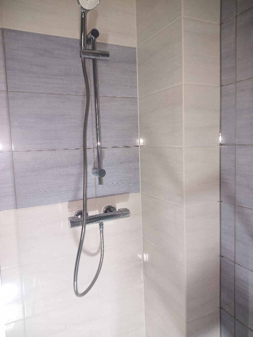 Chromowany zestaw natryskowy zamontowany na ścianie wyłożonej biało-szarymi płytkami w łazience. Widoczny mikser termostatyczny i deszczownica.