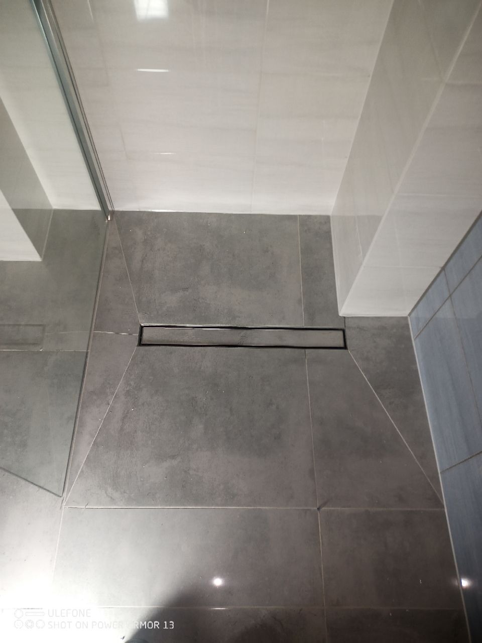 Wykończona łazienka z szarymi płytkami i odpływem liniowym. Widok z góry, minimalistyczny design. Fragment kabiny prysznicowej z elementami szkła.