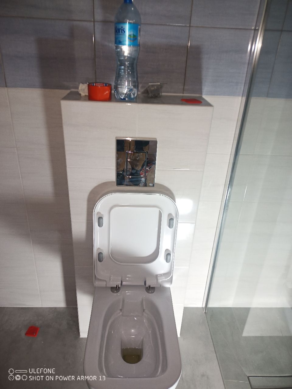 Podwieszana toaleta z otwartą klapą w wykończonej łazience, widoczna butelka wody na zabudowie stelaża. Szare płytki na ścianach i podłodze.