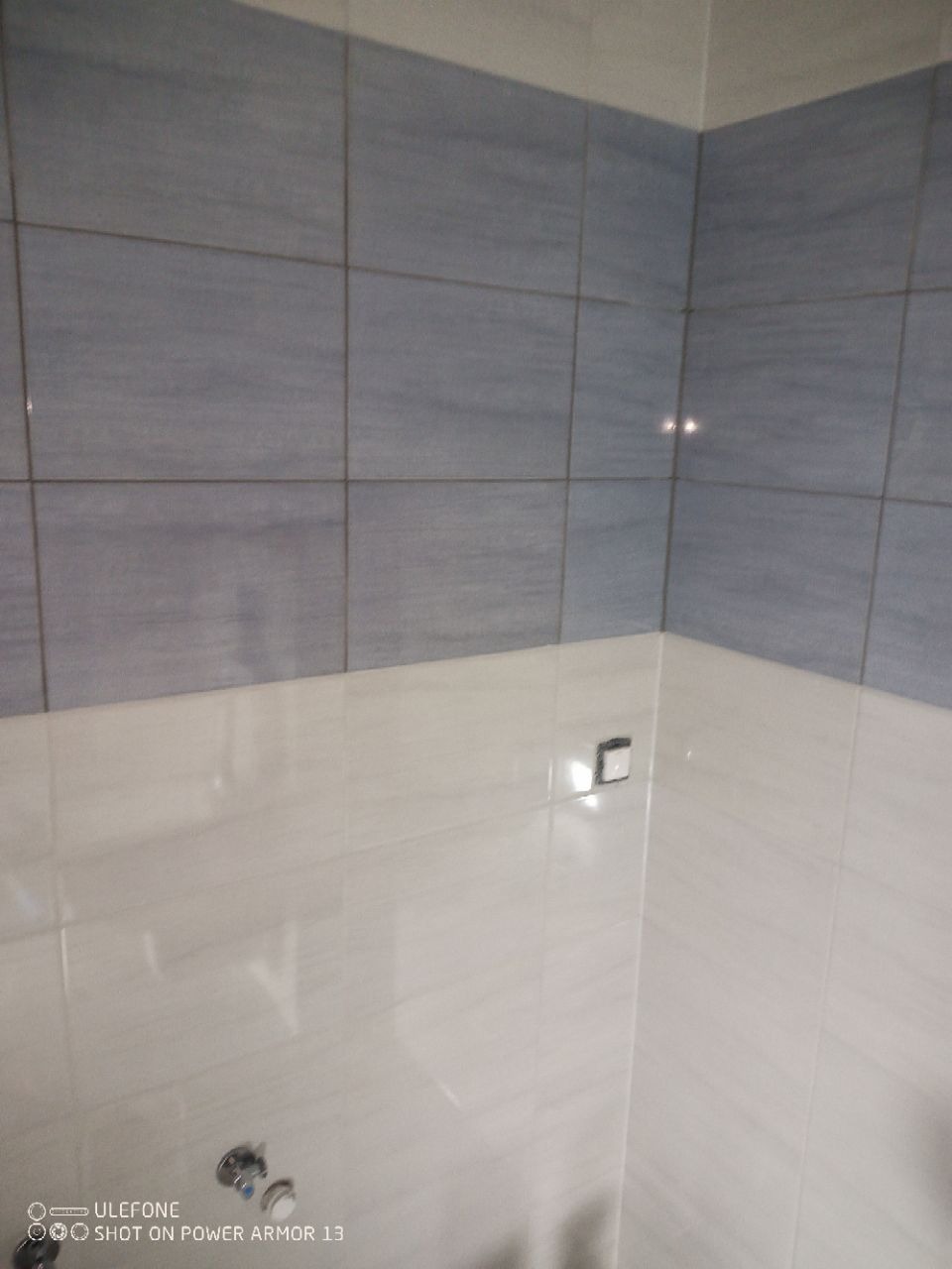 Wykończona ściana łazienki z dwukolorowymi płytkami: szare na górze i białe na dole. Widoczny chromowany zawór i uchwyt na słuchawkę prysznicową.