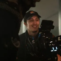 Uśmiechnięty kamerzysta w czapce z daszkiem, obsługujący profesjonalną kamerę filmową marki Miller, na planie zdjęciowym z rozmytym tłem i elementami oświetlenia.