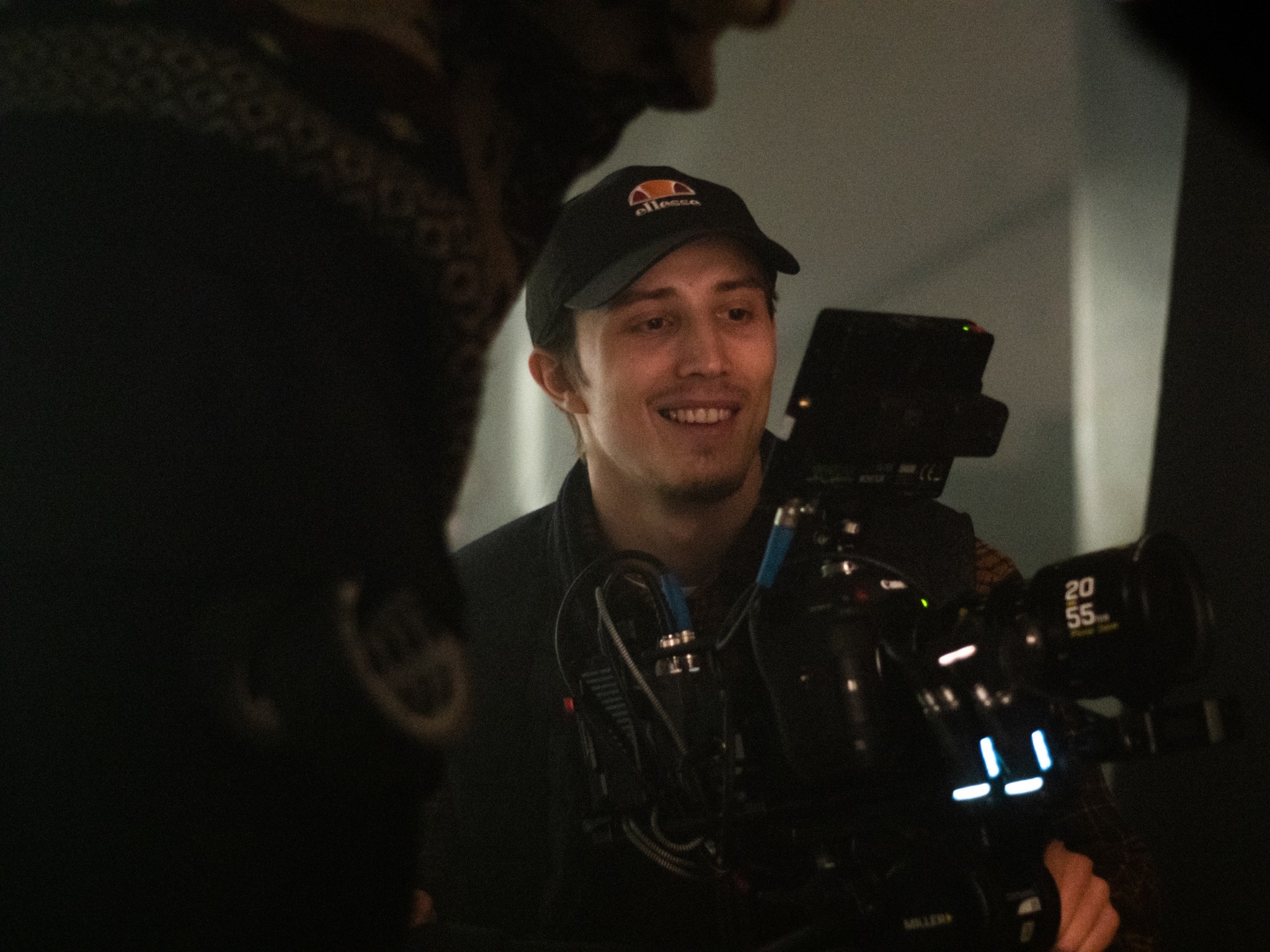 Uśmiechnięty kamerzysta w czapce z daszkiem, obsługujący profesjonalną kamerę filmową marki Miller, na planie zdjęciowym z rozmytym tłem i elementami oświetlenia.