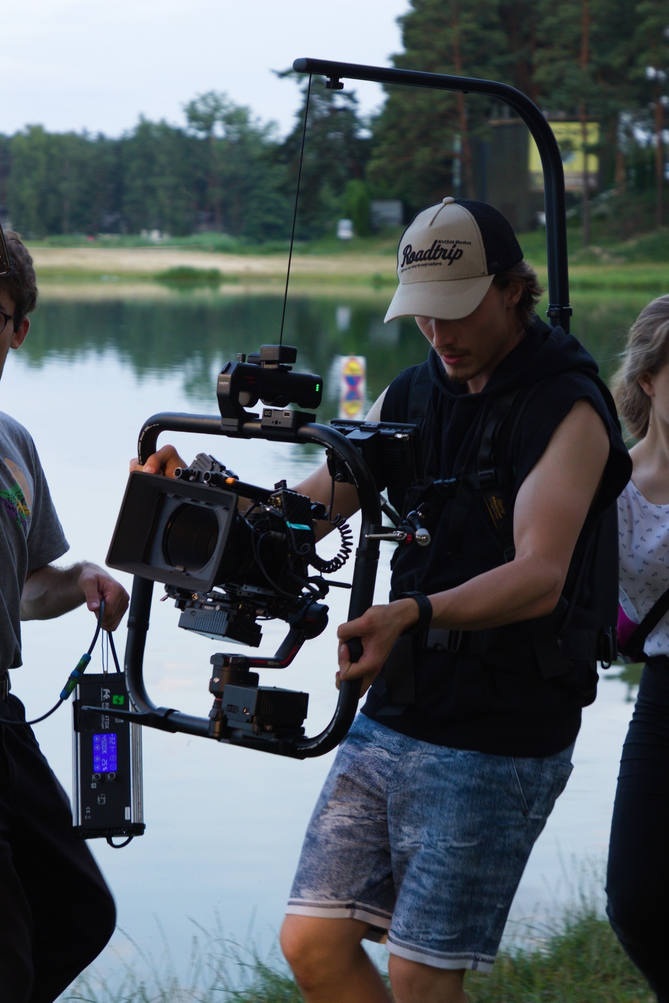 Ekipa filmowa kręci ujęcie nad jeziorem. Operator z gimbalem i kamerą, asystent z monitorem. W tle woda, las i lekko pochmurne niebo.