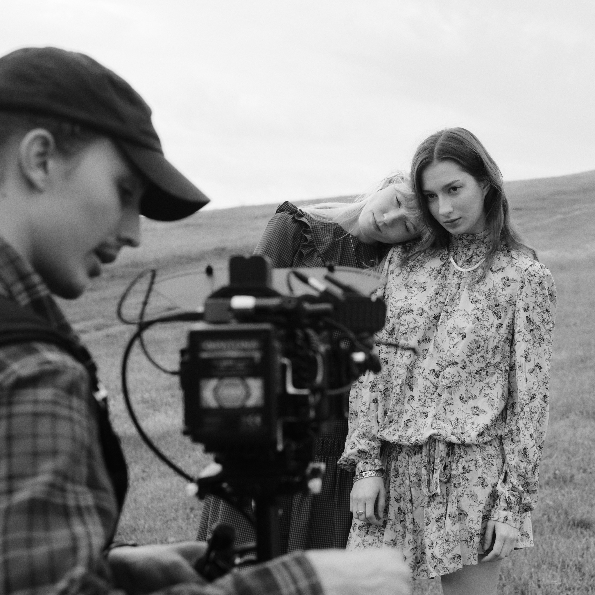 Czarno-białe ujęcie zza kamery na dwie modelki w sukienkach na tle wzgórza. Jedna opiera głowę na drugiej, patrząc w obiektyw. Uwagę zwraca szczegółowość faktur tkanin.