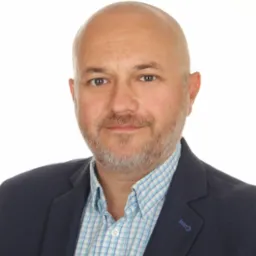 Portret biznesowy: łysiejący mężczyzna z brodą, w niebieskiej marynarce i koszuli w kratę, uśmiecha się na białym tle. Widoczne detale stroju.