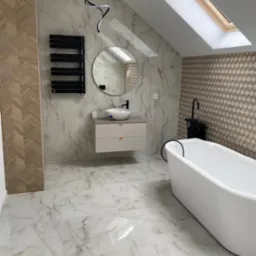 Nowoczesna łazienka na poddaszu z białą wanną wolnostojącą, okrągłym lustrem i marmurowymi płytkami. Uwagę przyciąga designerska lampa sufitowa i okno dachowe.