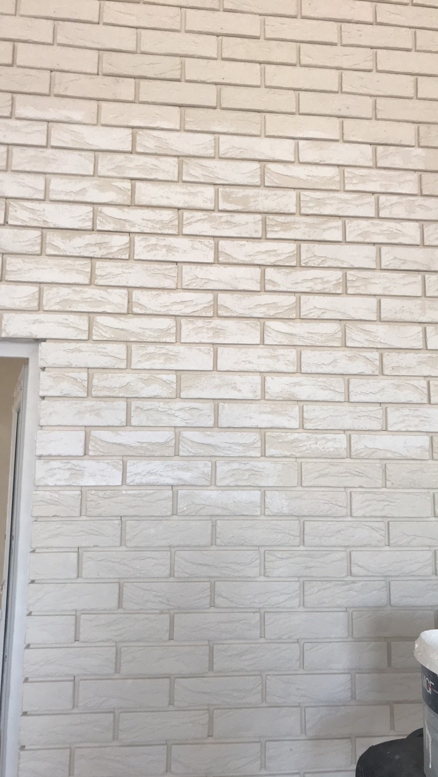 Ściana wyłożona białymi, dekoracyjnymi płytkami imitującymi cegłę, widoczne fugi. Fragment ościeżnicy po lewej.