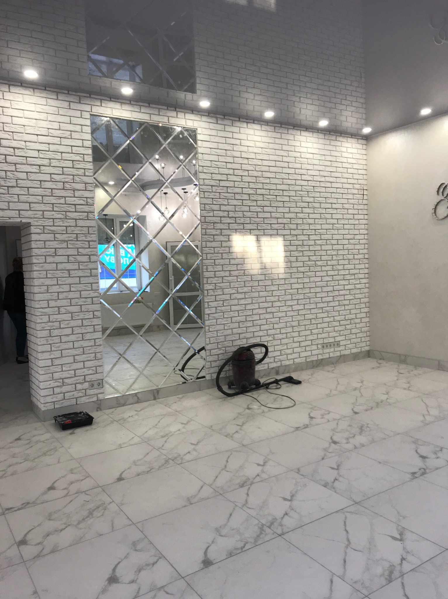 Wykończone wnętrze z lustrzaną mozaiką na ścianie z białej cegły, podświetlany sufit napinany i marmurowa podłoga. Widoczny odkurzacz i akcesoria malarskie.