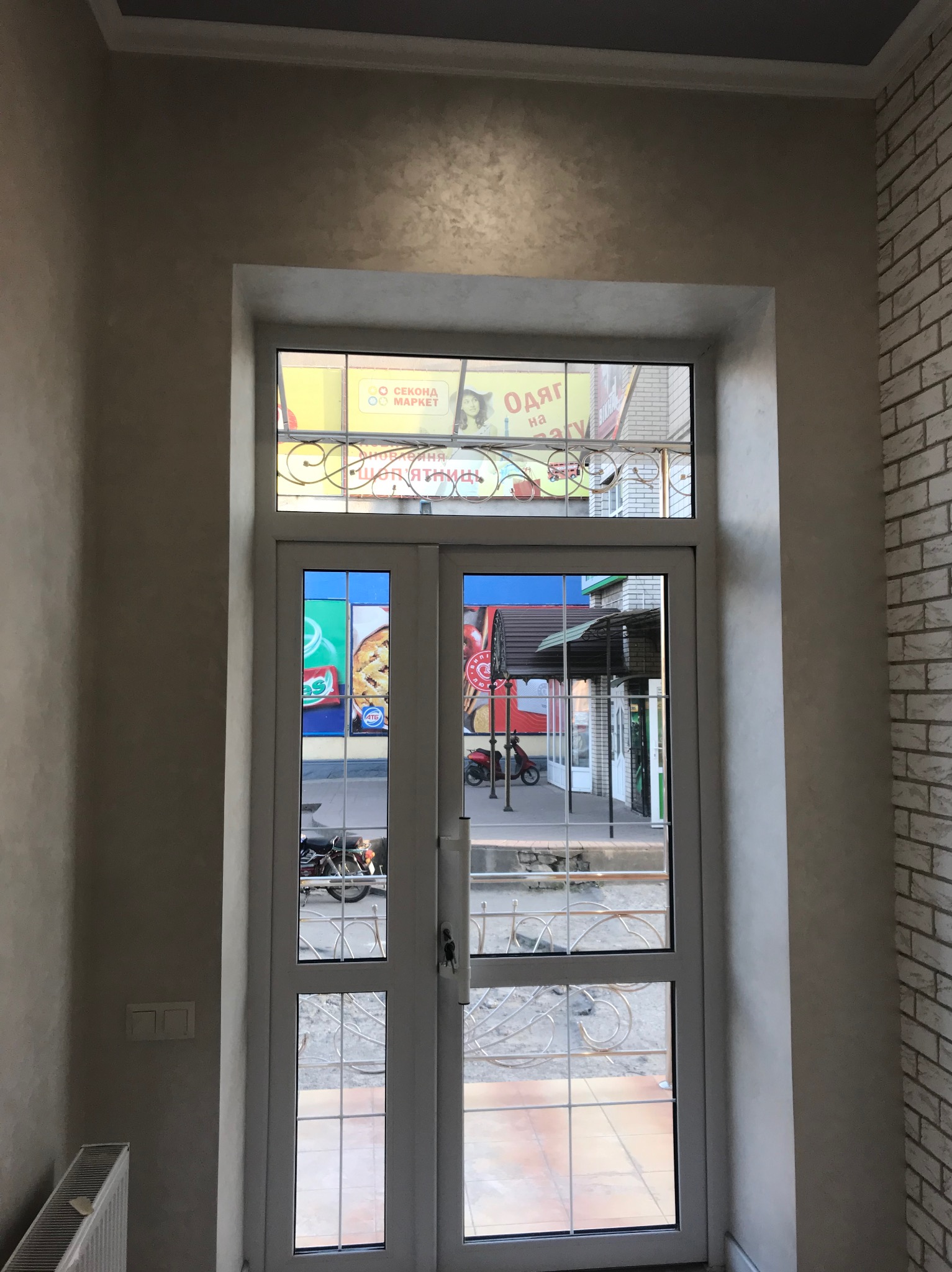 Białe, przeszklone drzwi balkonowe z ozdobną kratką, widok na ulicę, ściana z dekoracyjną fakturą i imitacją cegły. Fragment grzejnika.