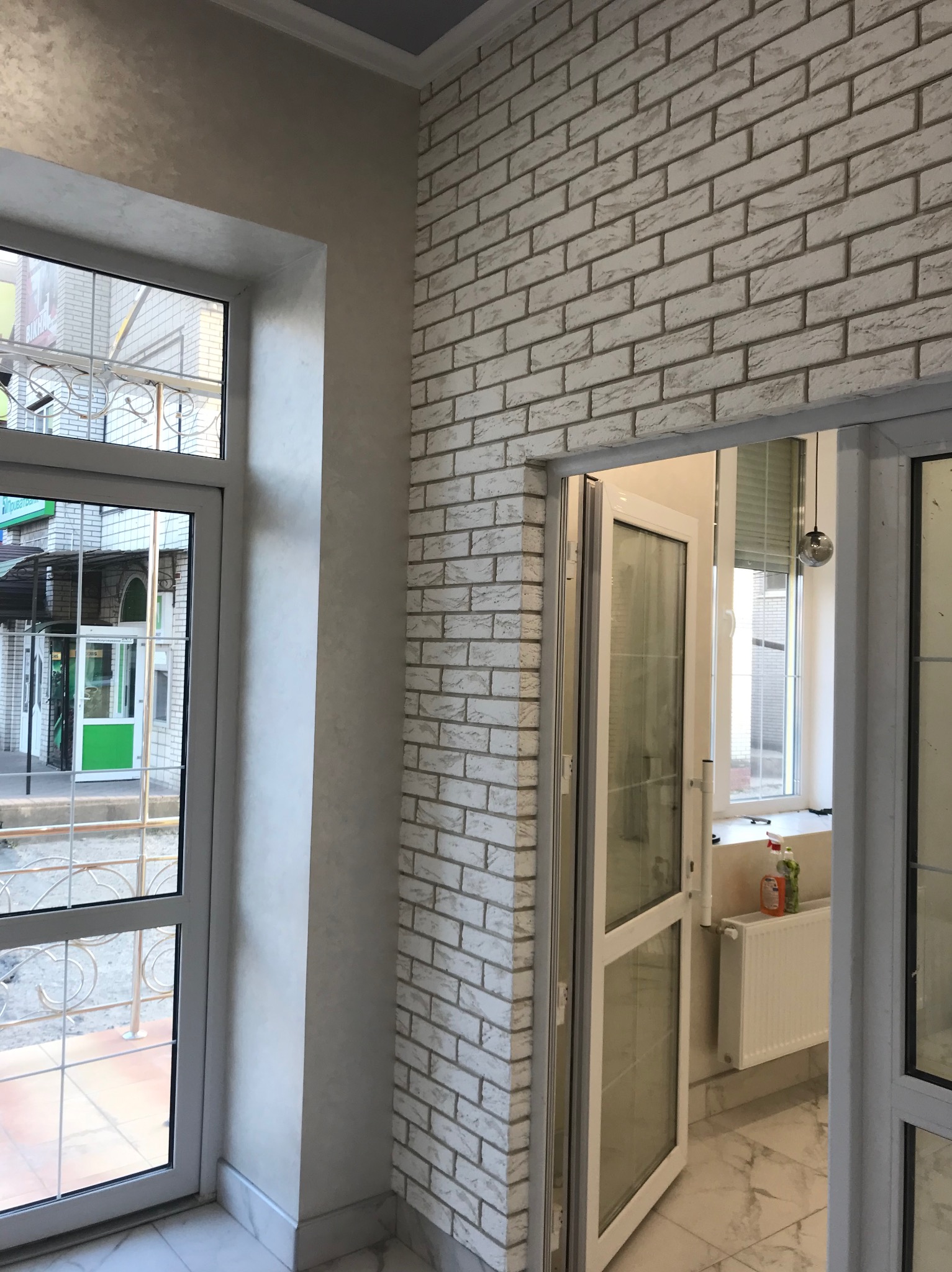 Wykończony fragment wnętrza z dekoracyjną ścianą z białej cegły i oknem z widokiem na zewnątrz, podkreślony neutralnym oświetleniem i marmurową podłogą.