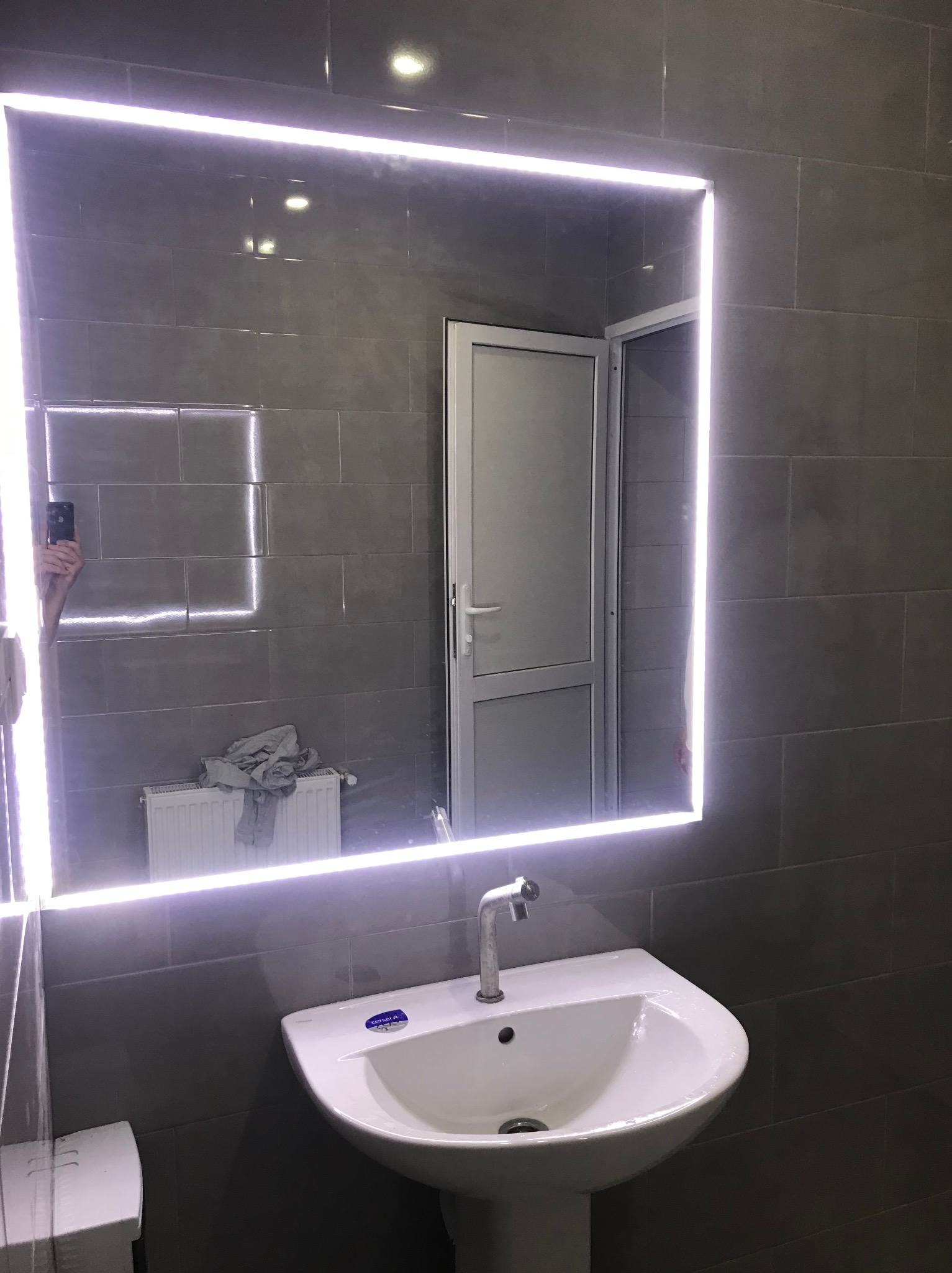Nowoczesna łazienka z podświetlanym lustrem LED nad umywalką na tle szarych płytek. W odbiciu widoczny fragment grzejnika i drzwi.