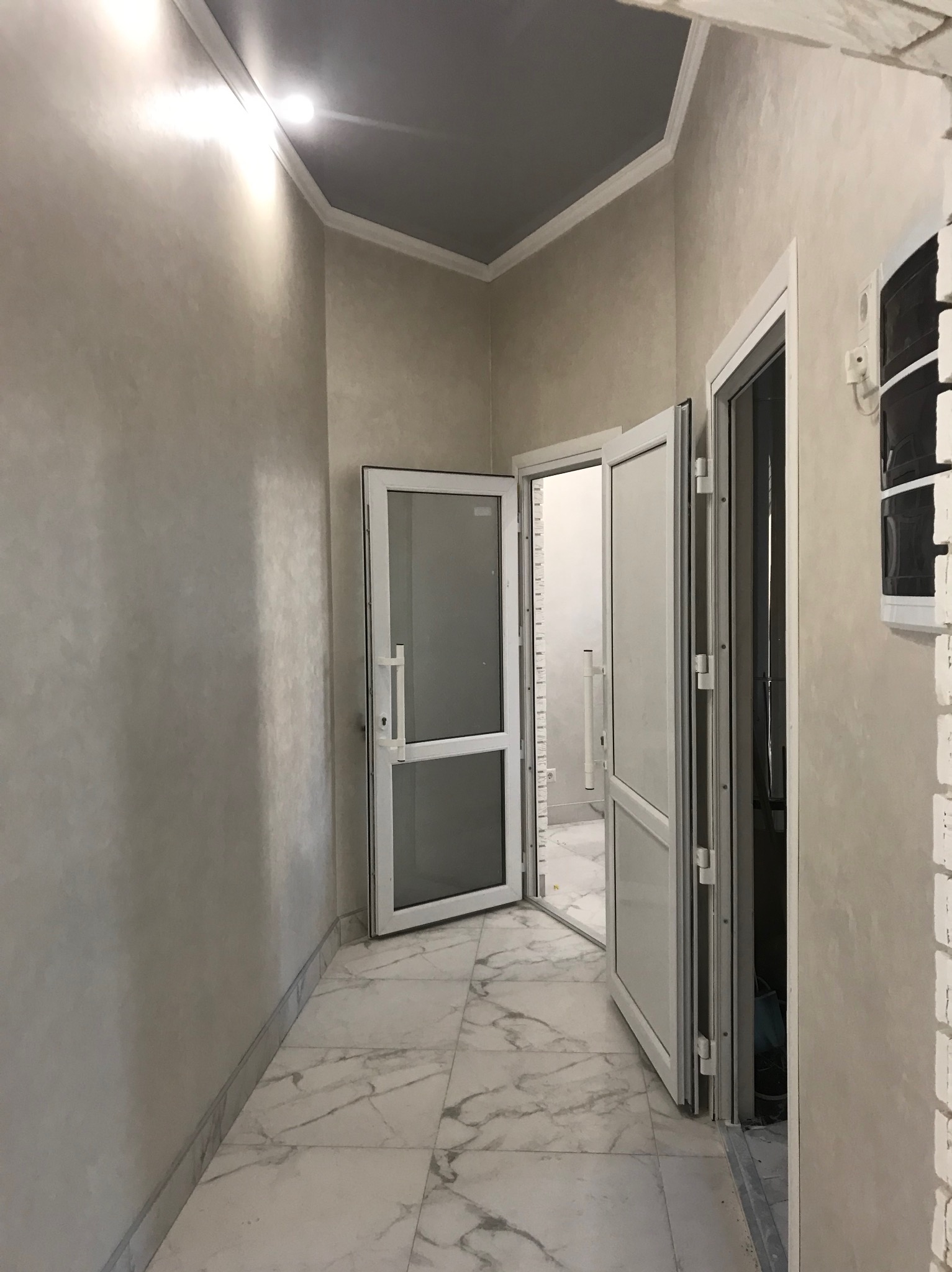 Wykończony korytarz z marmurową podłogą, pomalowanymi ścianami w odcieniach beżu i szarym sufitem. Widoczne białe drzwi z aluminiowymi ramami.