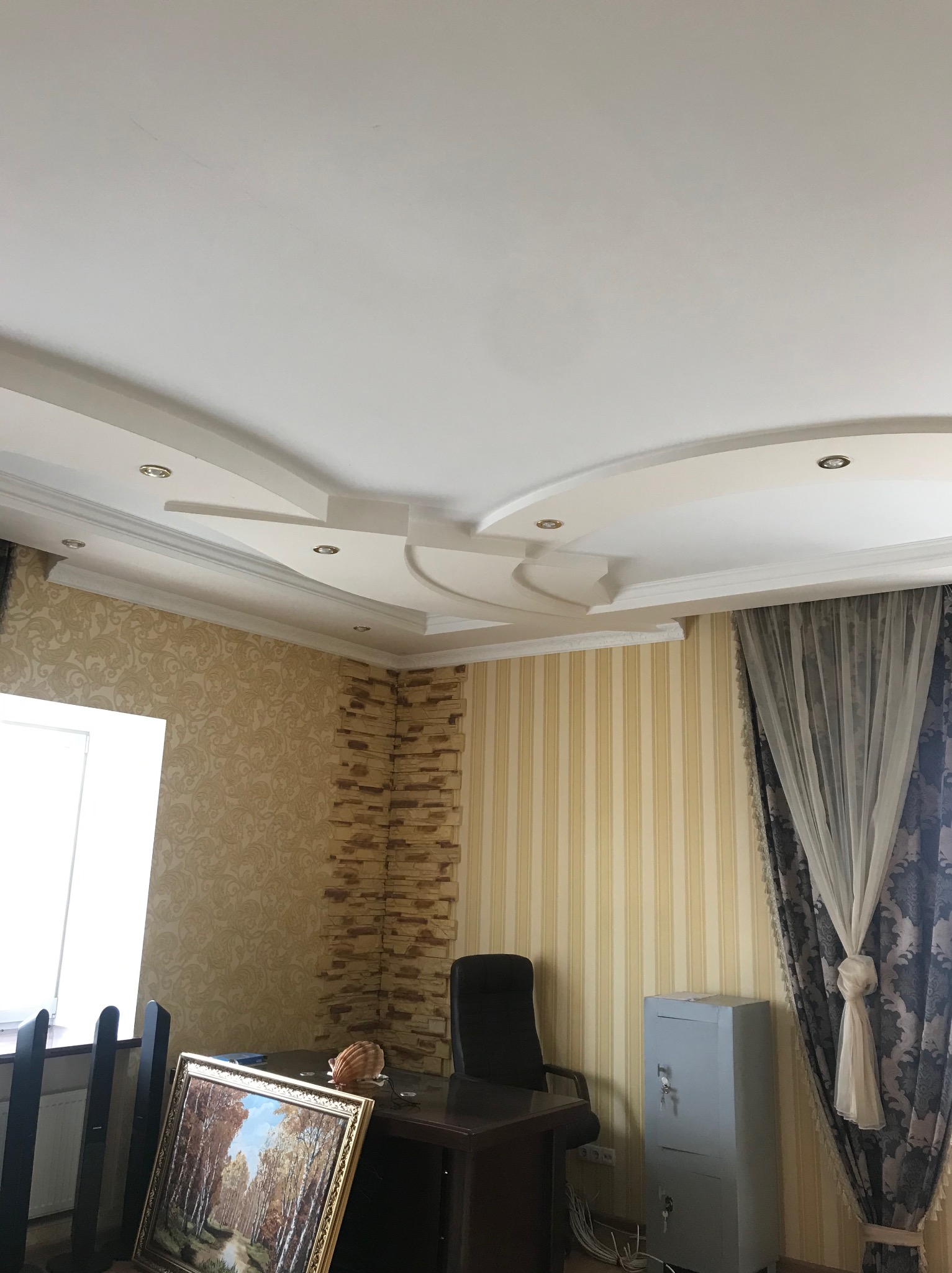 Dekoracyjny sufit podwieszany z oświetleniem punktowym w pokoju z tapetami w paski i wzorzystymi, ciężkimi zasłonami. Widoczny fragment biurka i obraz w złotej ramie.