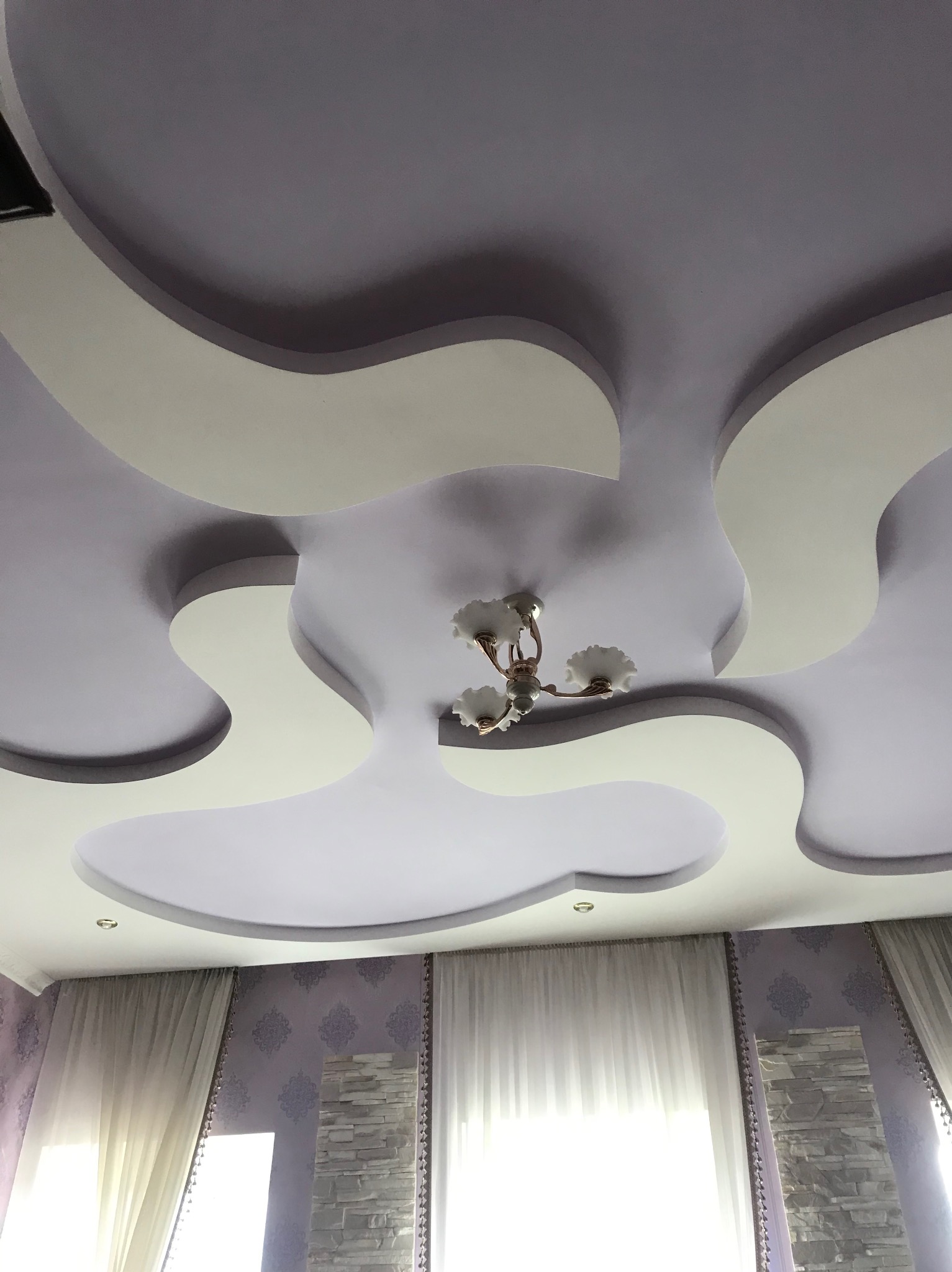 Wielopoziomowy, dekoracyjny sufit podwieszany w odcieniach bieli i fioletu z nowoczesnym oświetleniem, widok z dołu. W tle ściana z wzorzystą tapetą i zasłony.