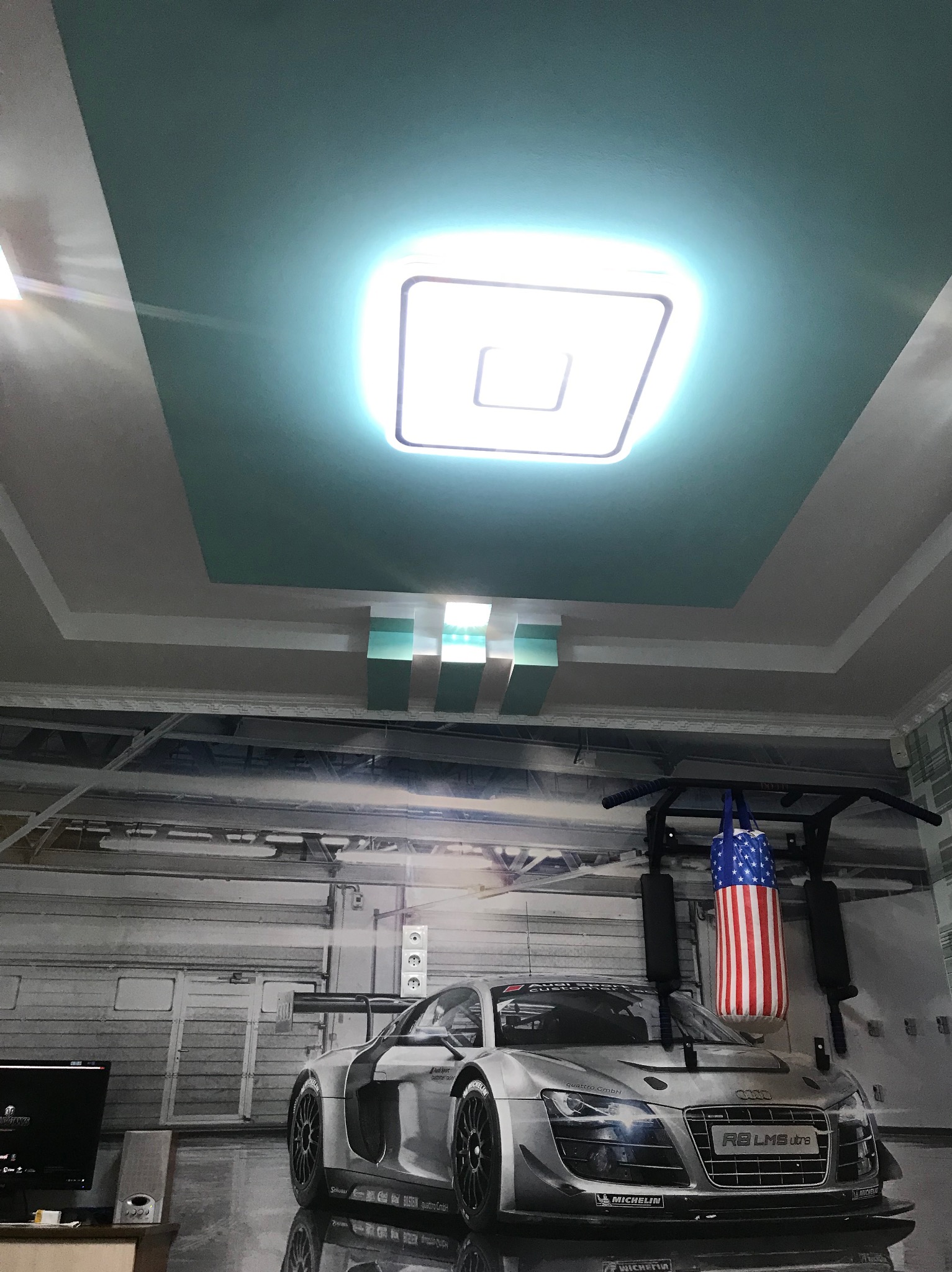 Nietypowy sufit podwieszany z wbudowanym oświetleniem LED i dekoracyjnymi elementami. Na ścianie fototapeta z motywem sportowego samochodu i workiem bokserskim.