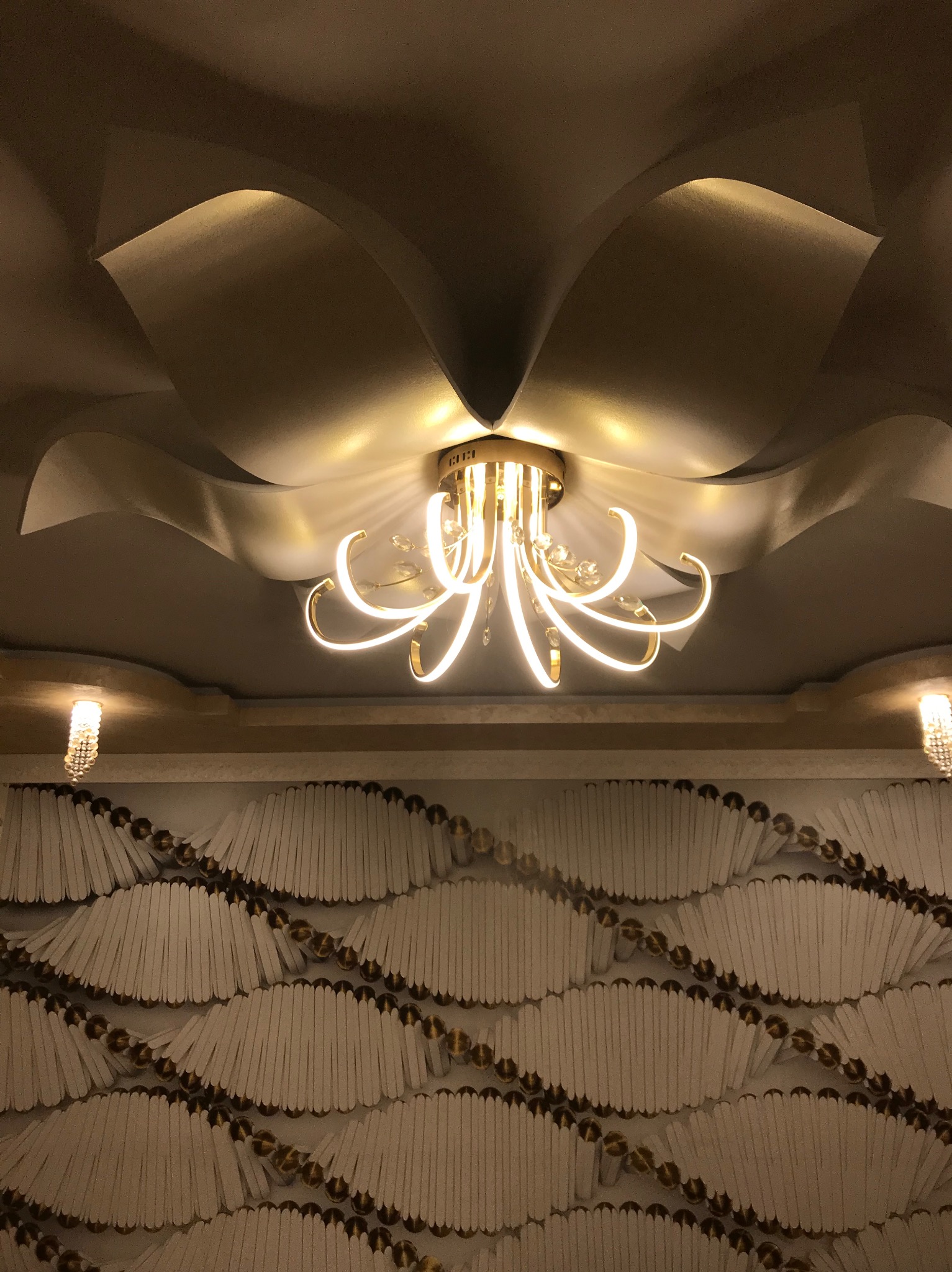 Dekoracyjny sufit z podwieszanymi elementami i nowoczesnym żyrandolem z kryształkami, oświetlony ciepłym światłem.