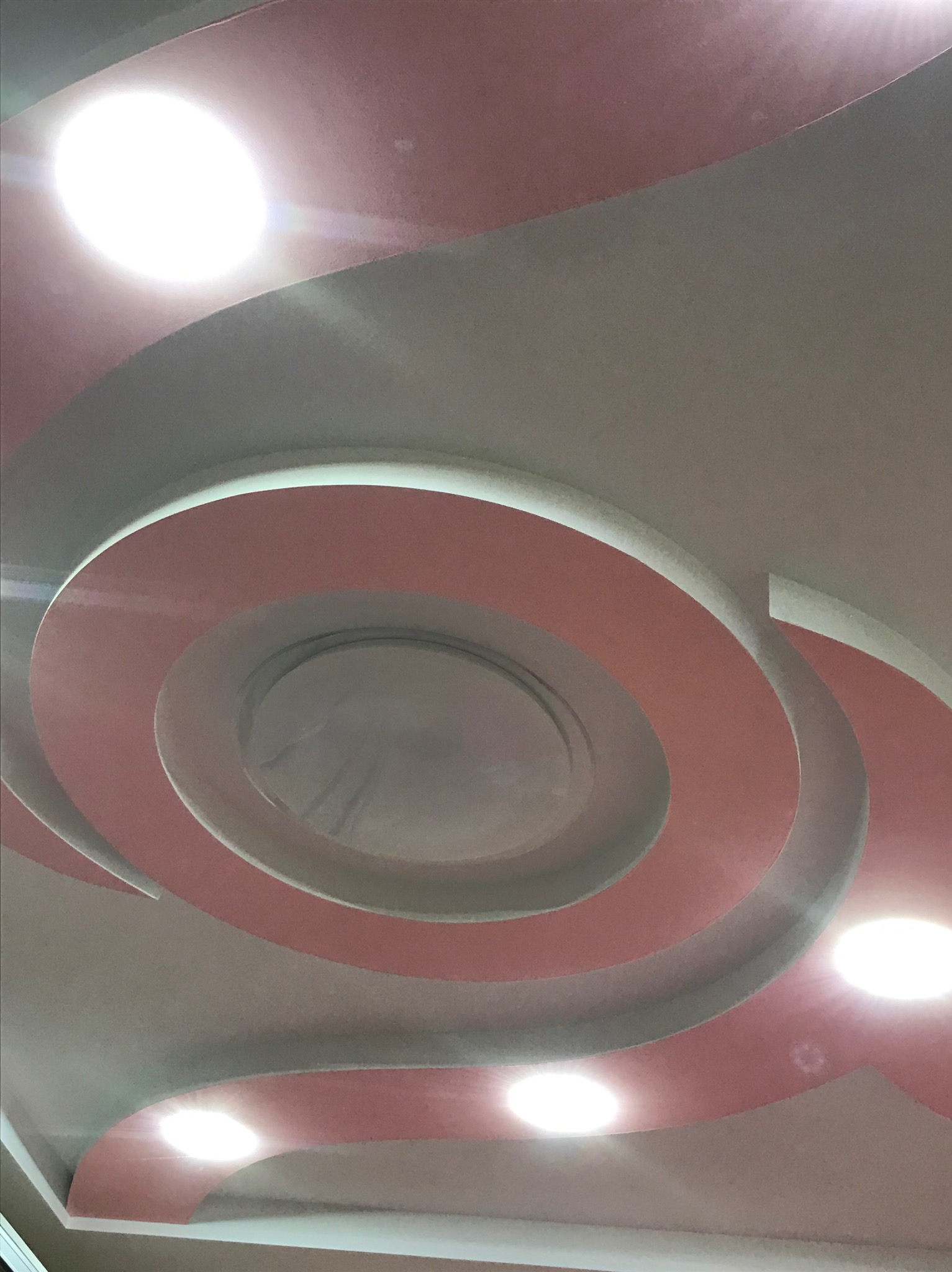 Dekoracyjny sufit podwieszany z okrągłymi elementami w odcieniach różu i szarości, oświetlony punktowymi lampami, tworzący nowoczesny design wnętrza.