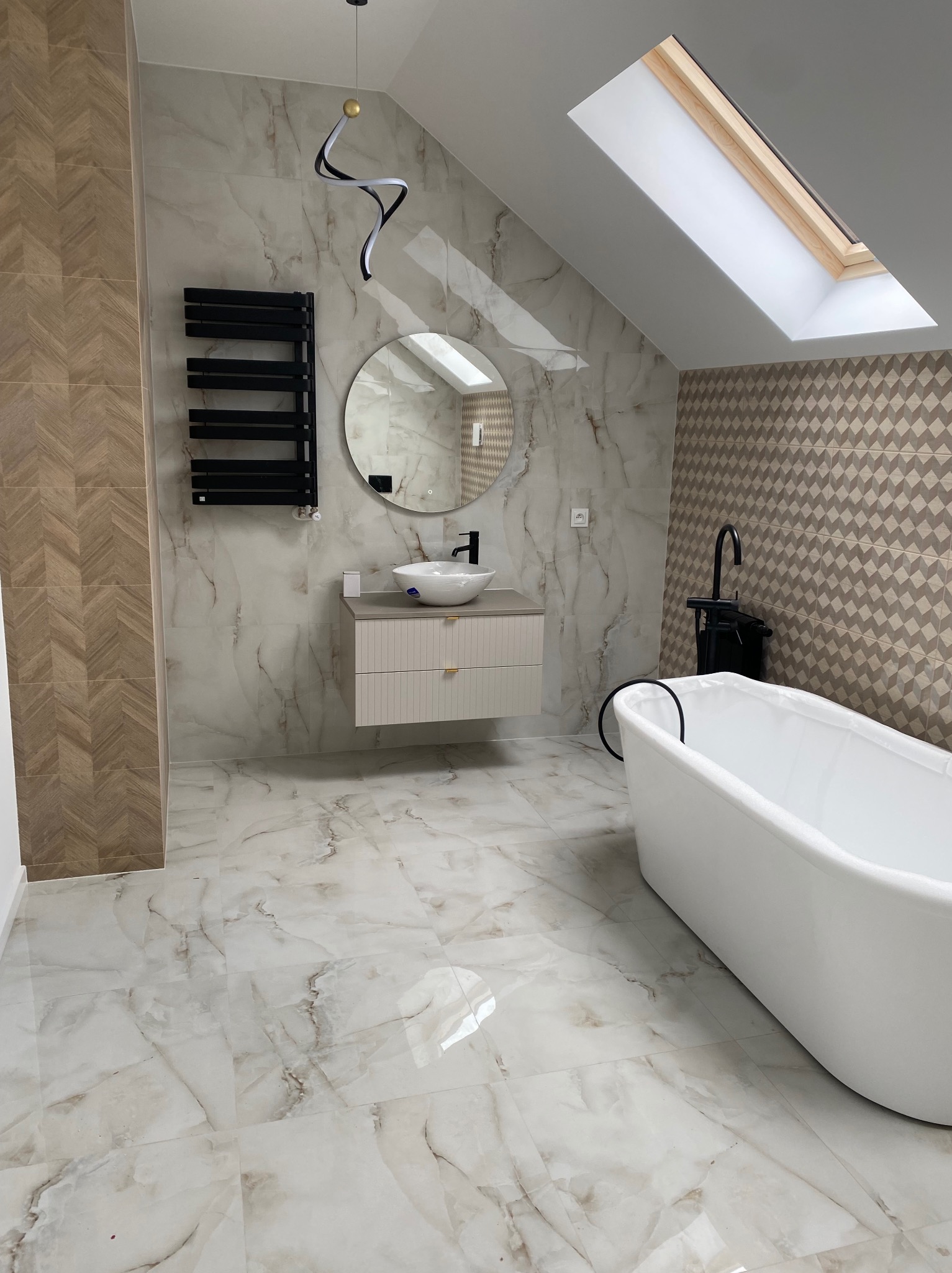 Nowoczesna łazienka na poddaszu z białą wanną wolnostojącą, okrągłym lustrem i marmurowymi płytkami. Uwagę przyciąga designerska lampa sufitowa i okno dachowe.